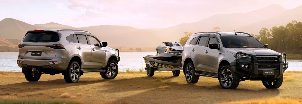 A silver Isuzu MU-X Tour Mate, equipped with the factory bull bar, towing a trailer along a dusty dirt road next to a large lake