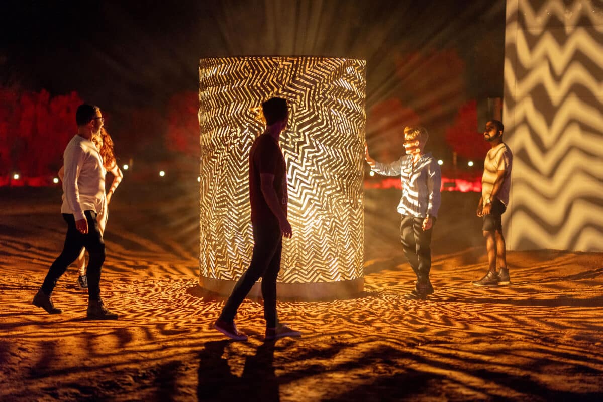 Visitors walk through the 'Transforming Light & Country' installation at Parrtjima, featuring multiple large arches illuminated with white and blue lights on the red earth at night.