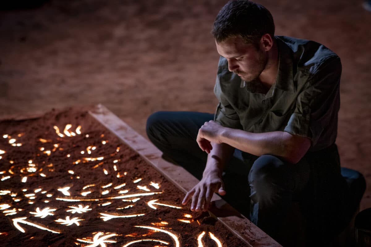 A man crouches to interact with an illuminated sandpainting installation at Parrtjima, his hand gently touching the red sand where glowing white patterns shine through