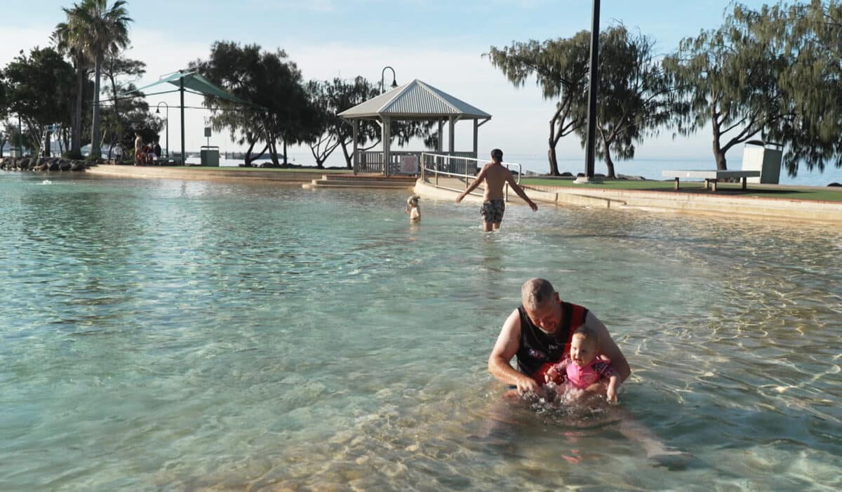 A man sitting in the public wading pool at Scarborough, qld, holds his young smiling baby girl as she splashes in the shallows