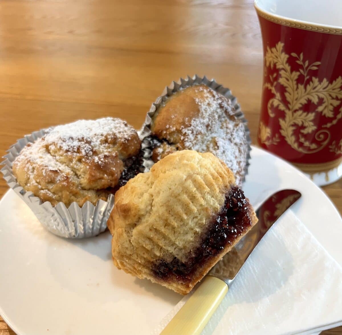 Two and a half easy bake jam doughnut muffins rest on a white plate with a napkin and a butter now. There is a red cup with a gold leaf pattern in the background. Recipe below. Uses few ingredients. Perfect for caravanners