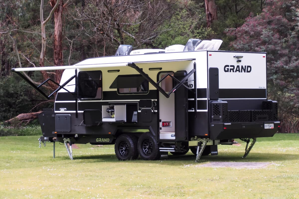 external view of the Grand Caravans 2025 Peak caravan on green grass in front of treed location. Note the fully extended Carefree electric awning