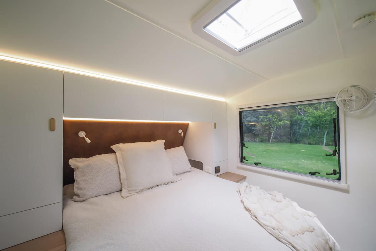 Caravan interior showing a queensize bed surrounded by white wardrobes adjacent a large window.