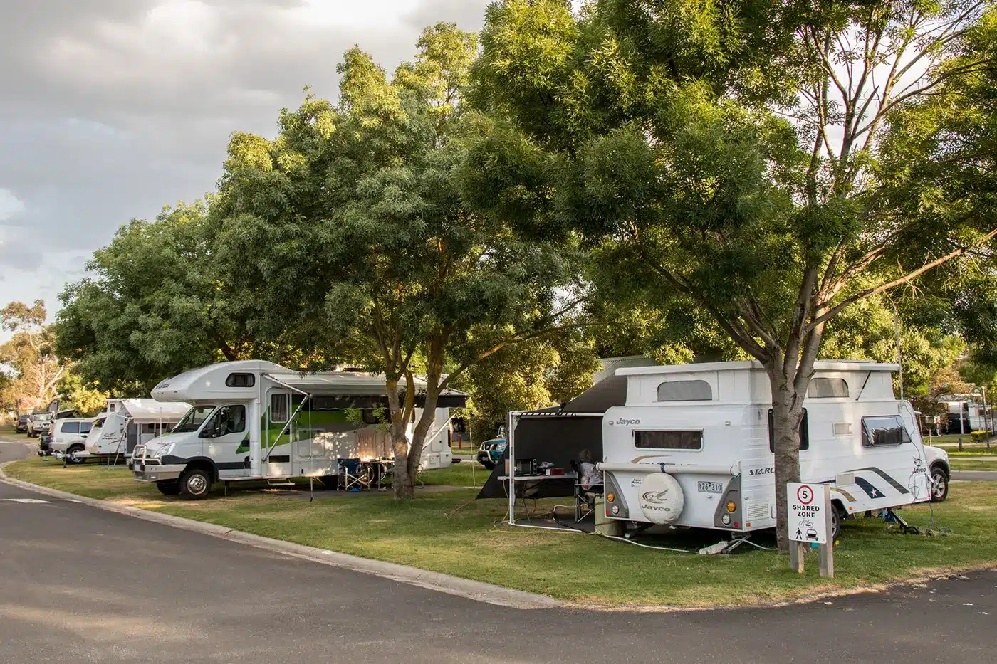 Caravans and motorhomes set up on powered grass sites in a holiday park with trees and shade