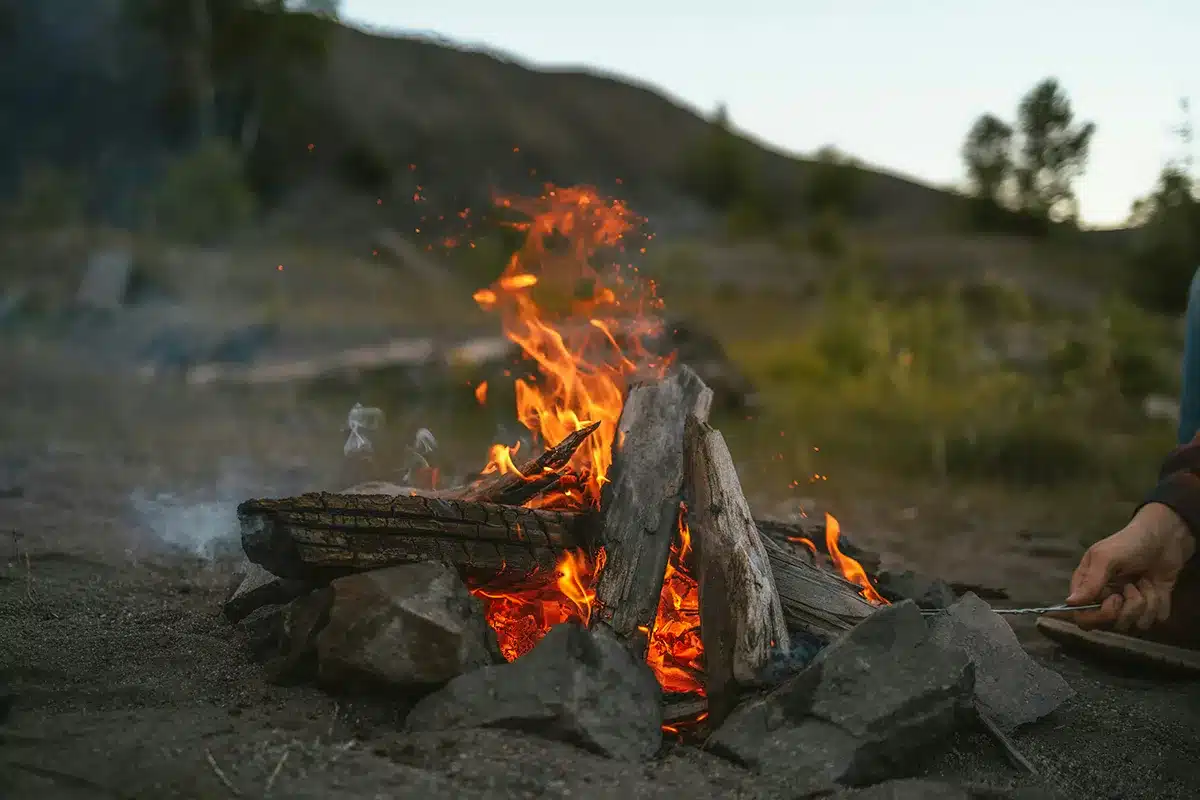 Campfire burning with open flames and sparks at a campsite at dusk, with rocks forming a fire ring and bushland hills in the background