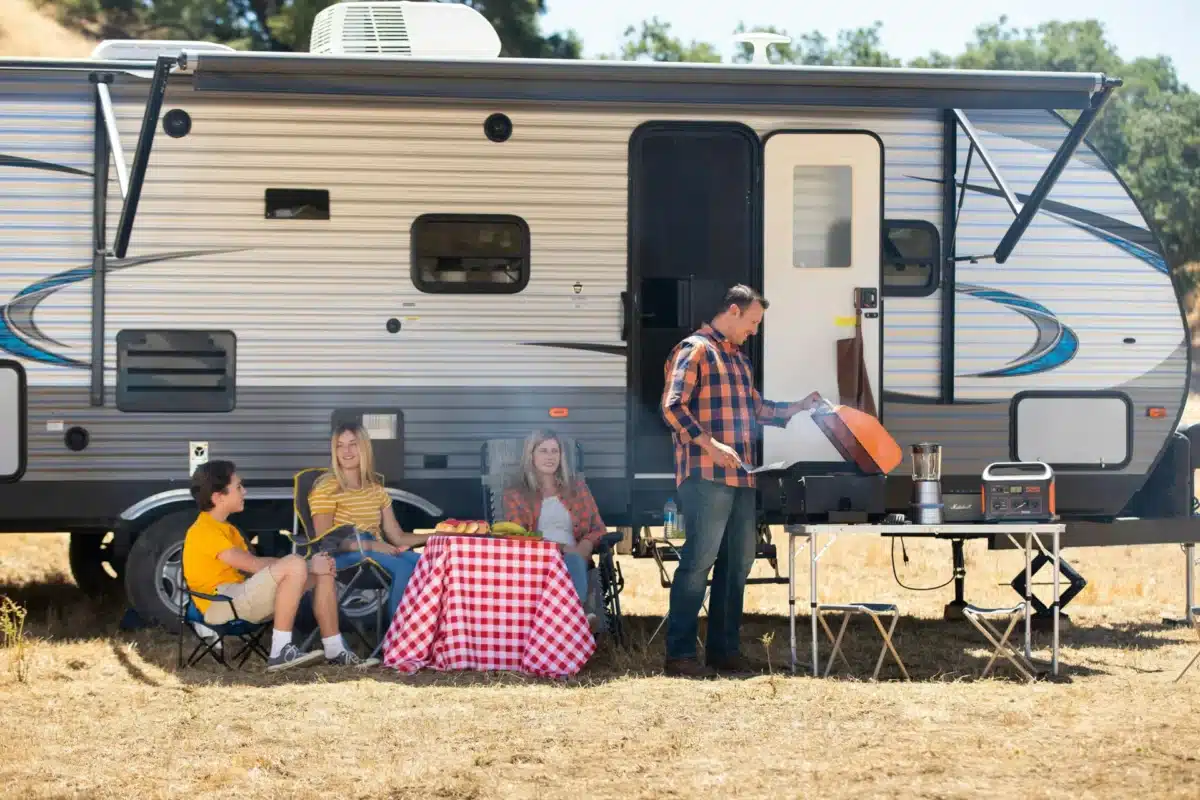 Family of four cooking and relaxing outside their caravan at a campsite, with a dad grilling at a portable camp table and kids seated around a picnic table with fresh fruit